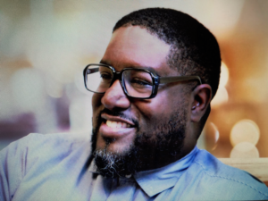 Headshot of Malik Singleton shows me smiling and wearing eyeglasses.